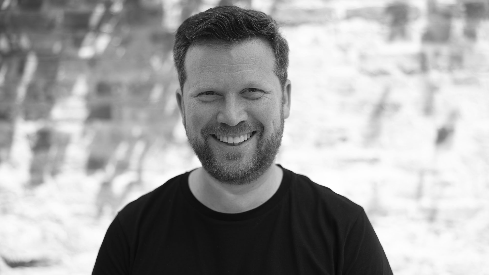 Ein Mann mit Vollbart lächelt offen in die Kamera. Er trägt ein schwarzes T-Shirt. Im Hintergrund kann man eine Mauer erahnen.